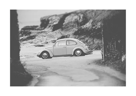 Zwart-witfoto van een klassieke Volkswagen Kever bij een strand met rotsen