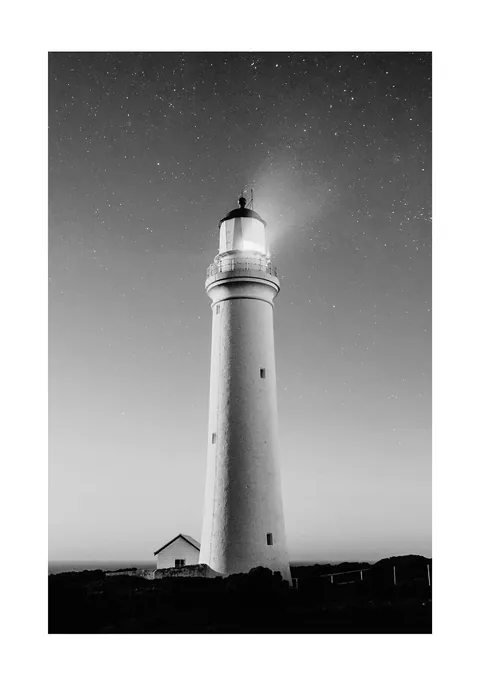 Zwart-witfoto van een verlichte witte vuurtoren onder een heldere sterrenhemel.